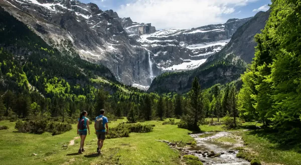 Dos excursionistas caminando por un valle verde de los Pirineos con montañas nevadas y cascada al fondo.