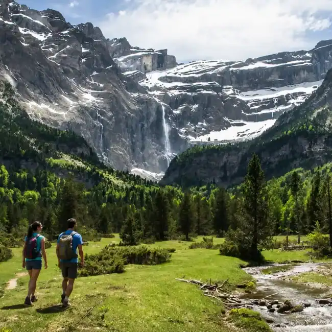 Dos excursionistas caminando por un valle verde de los Pirineos con montañas nevadas y cascada al fondo.