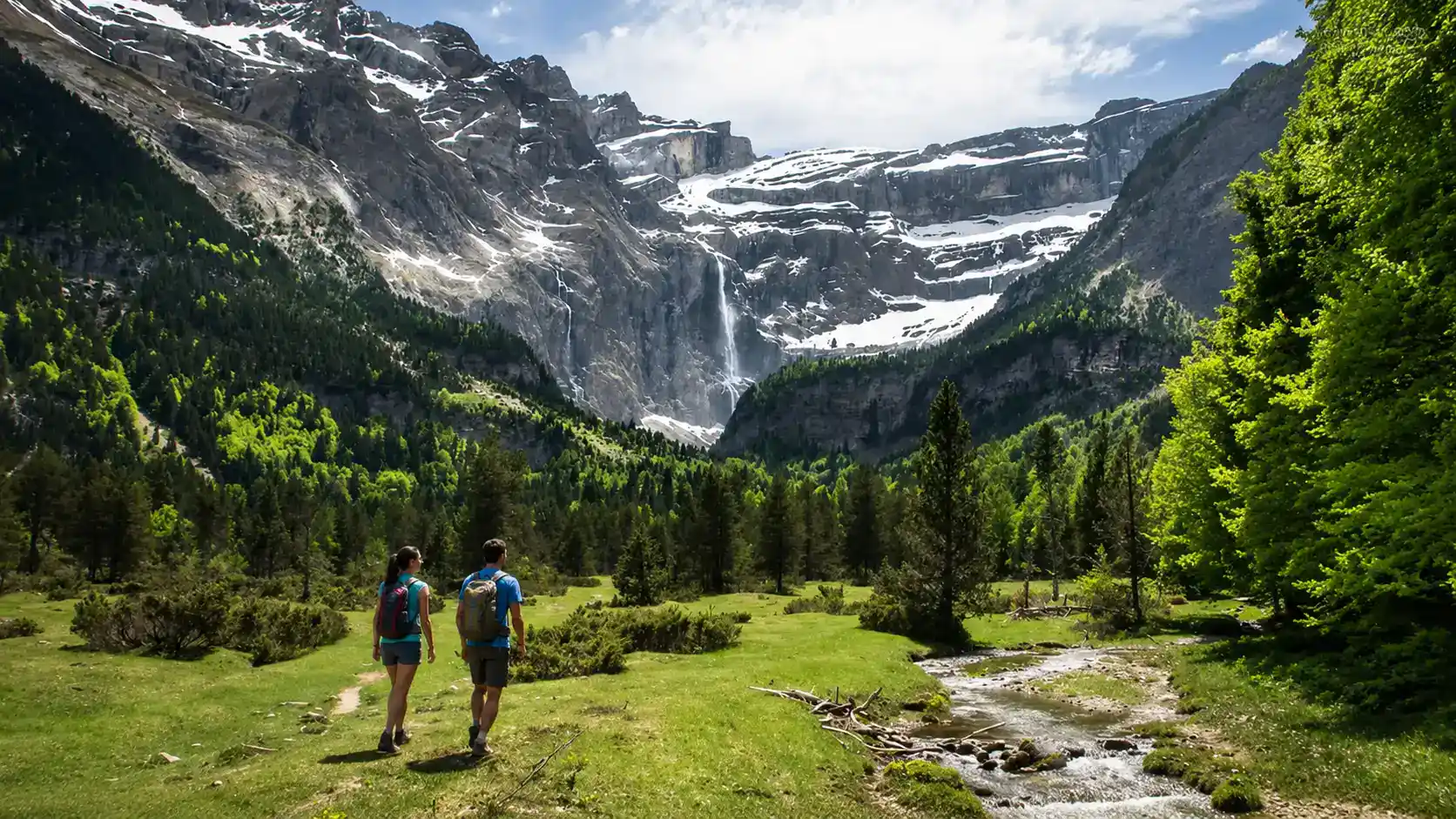 Dos excursionistas caminando por un valle verde de los Pirineos con montañas nevadas y cascada al fondo.